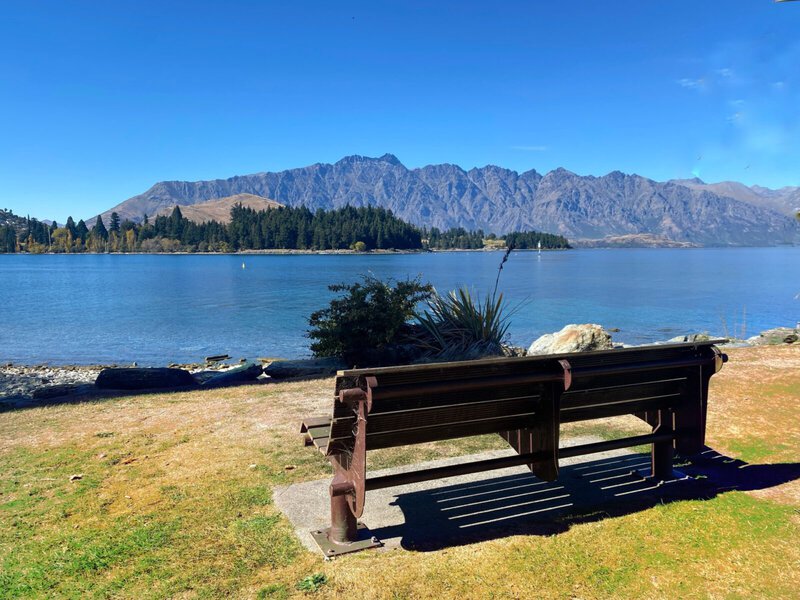 Eine Holzbank mit Blick auf den Lake Wakatipu in Queenstown, Neuseeland, umgeben von Bergen und klarem blauen Himmel.