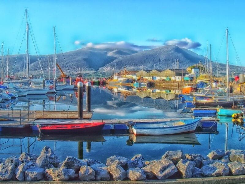 Malerischer Hafen mit bunten Booten, ruhigem Wasser und Bergen im Hintergrund unter blauem Himmel.