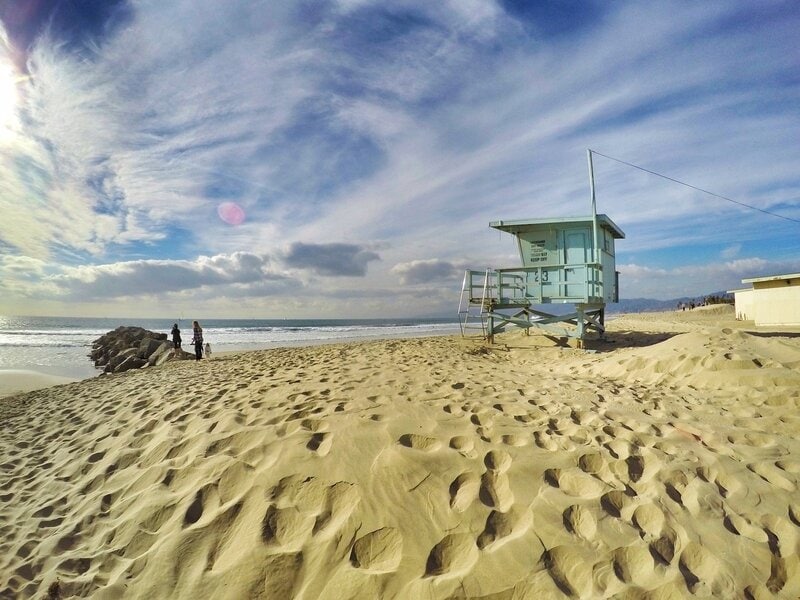 Ein Sandstrand in Kalifornien mit einem blauen Rettungsschwimmerturm, dem Meer und einem dramatischen Himmel im Hintergrund.