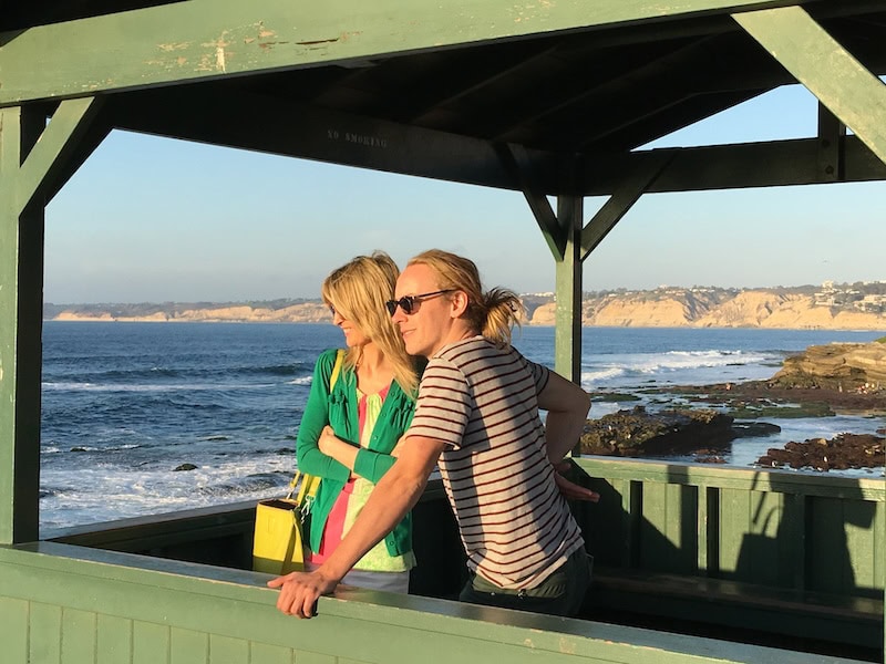 Young man and woman standing in a green wooden lookout shelter overlooking the rocky shoreline and waves at a San Diego beach on a clear sunny afternoon.