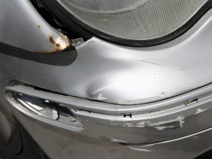 Damaged car with a noticeable bumper dent, scratches, and rust near the headlight on a silver surface.