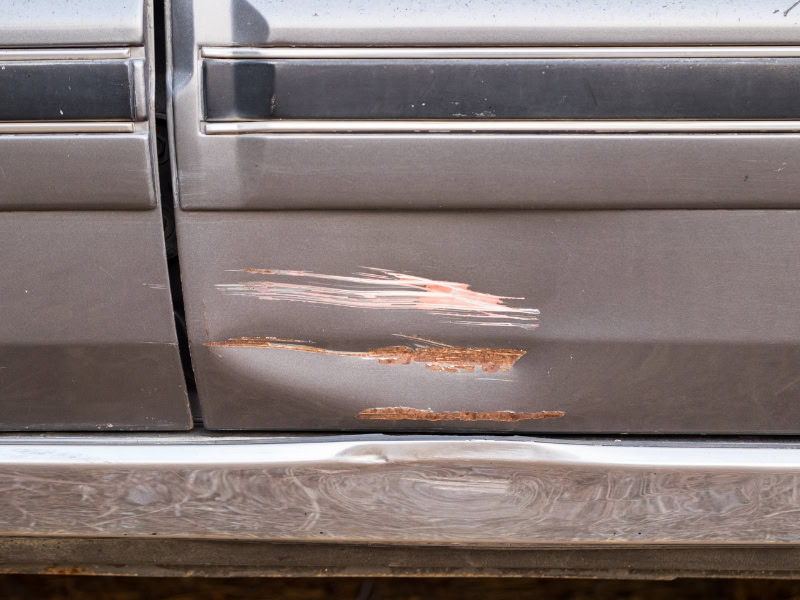 Close-up of a car scratch causing rust on a grey car door, showing damage to the paintwork.