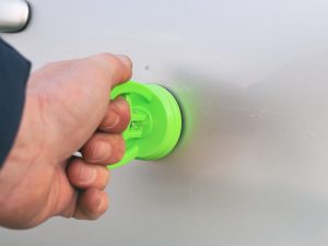 A hand uses a green suction cup tool for DIY hail damage repair on a car's panel.