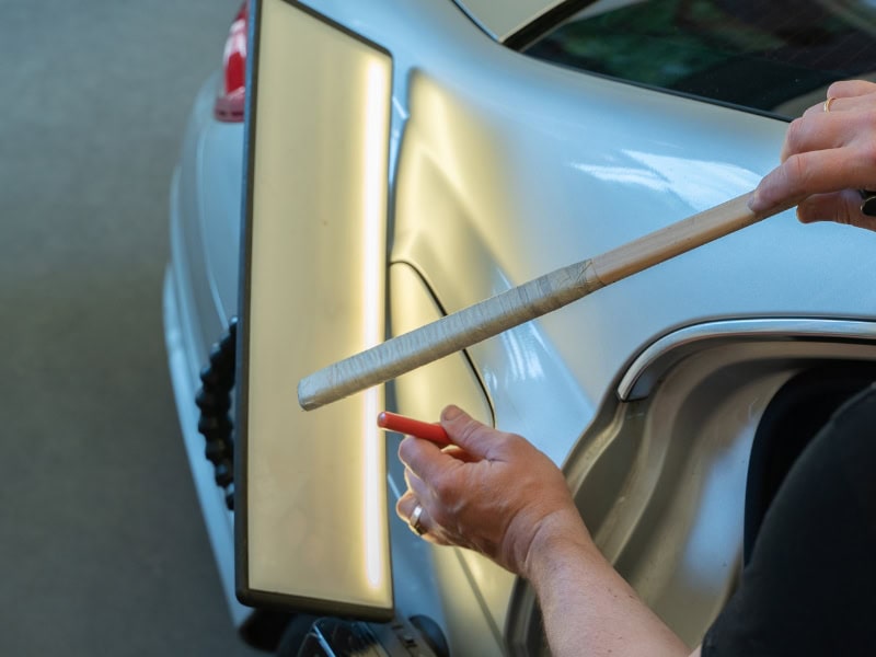 Local car dent repair technician using reflection board and tool for panel dent assessment. 