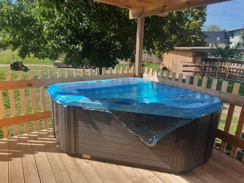 Entspannender Whirlpool auf Holzterrasse im Grünen, ideal für Wellness und Erholung, eingewickelt in Schutzplane, umgeben von Natur, perfekt für Wellnesshof-Raja Gäste in Deutschland.