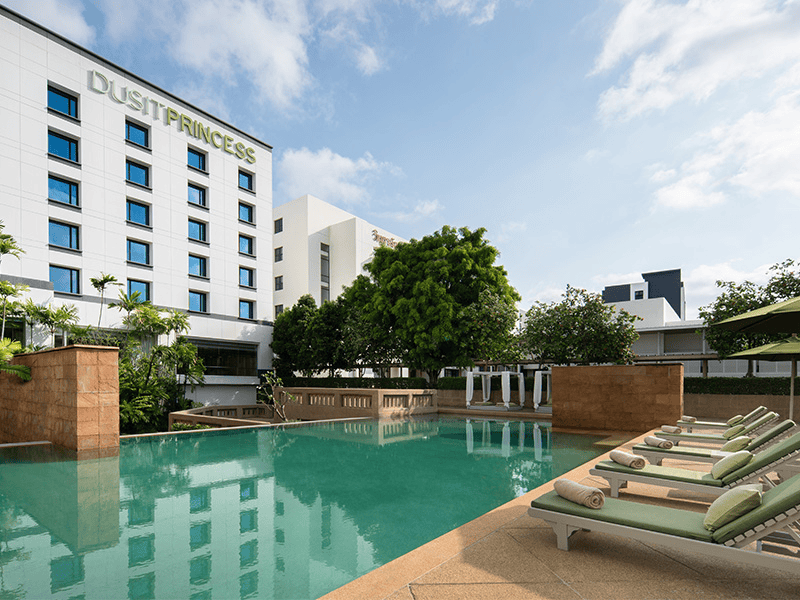 A clean, modern hotel building named "Dusit Princess" with a clear outdoor swimming pool and sun loungers lined up beside it.