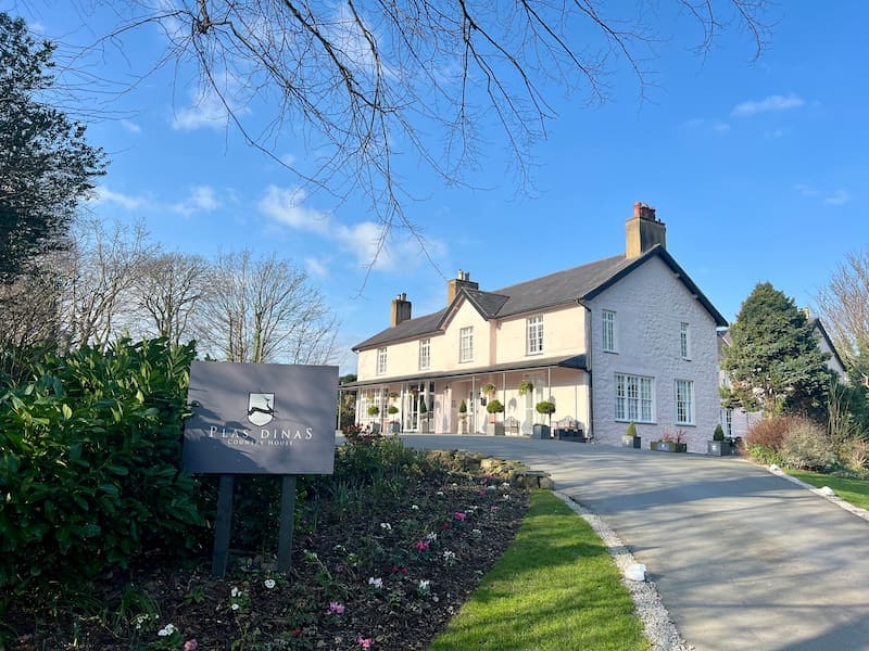 The exterior of Plas Dinas Country House, a historic cream-colored estate with a wraparound porch, nestled among manicured gardens. A sign with the property's name is positioned in the foreground, while a clear blue sky and lush trees create a picturesque setting. The scene feels peaceful and welcoming.