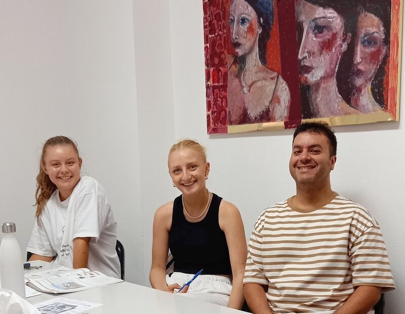 Drei Sprachschüler sitzen lächelnd an einem Tisch mit Lernmaterialien in einem Klassenzimmer mit moderner Kunst an der Wand.