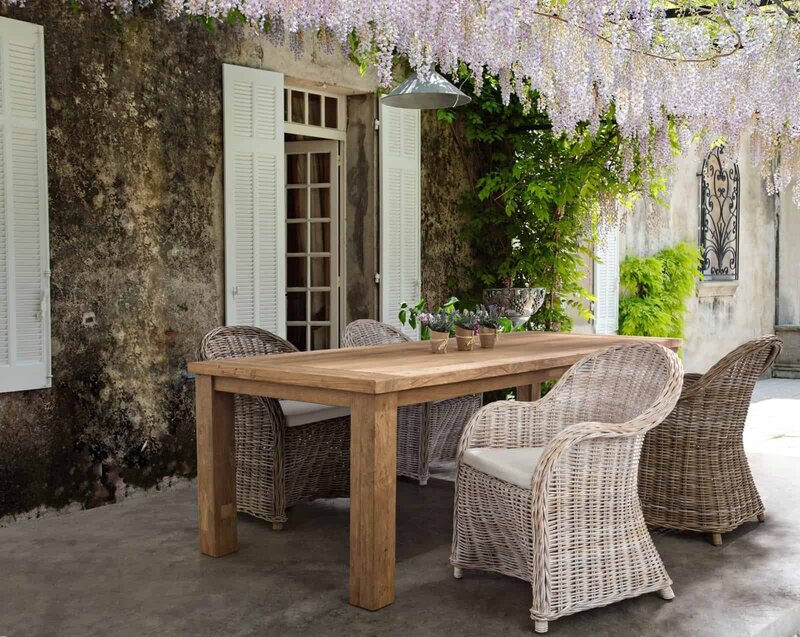 Assenza, ambiente esterno con tavolo in legno e sedie in vimini sotto glicine in fiore, atmosfera rilassante.