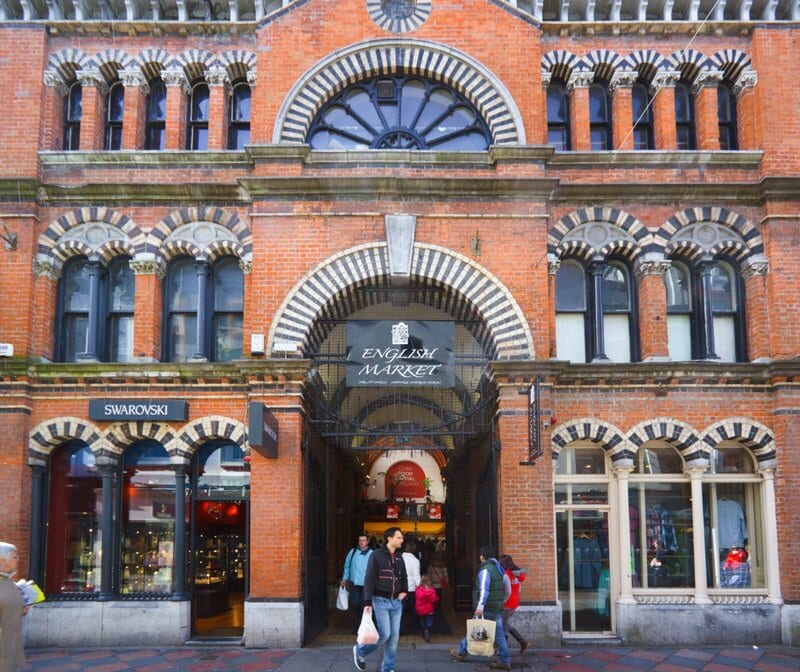 Historischer Eingang zum English Market in Cork, Irland, mit roten Backsteinbögen und Passanten.