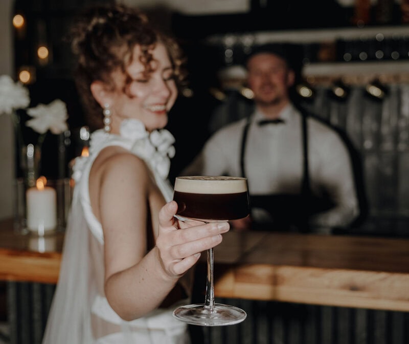 Eine Braut in einem weißen Kleid lächelt und hält ein Cocktailglas nach vorne in den Fokus, mit einem verschwommenen Barkeeper hinter einer hölzernen Bar - die perfekte Inspiration für Ihre Hochzeitsplanung Bayern in dieser warm beleuchteten Umgebung.