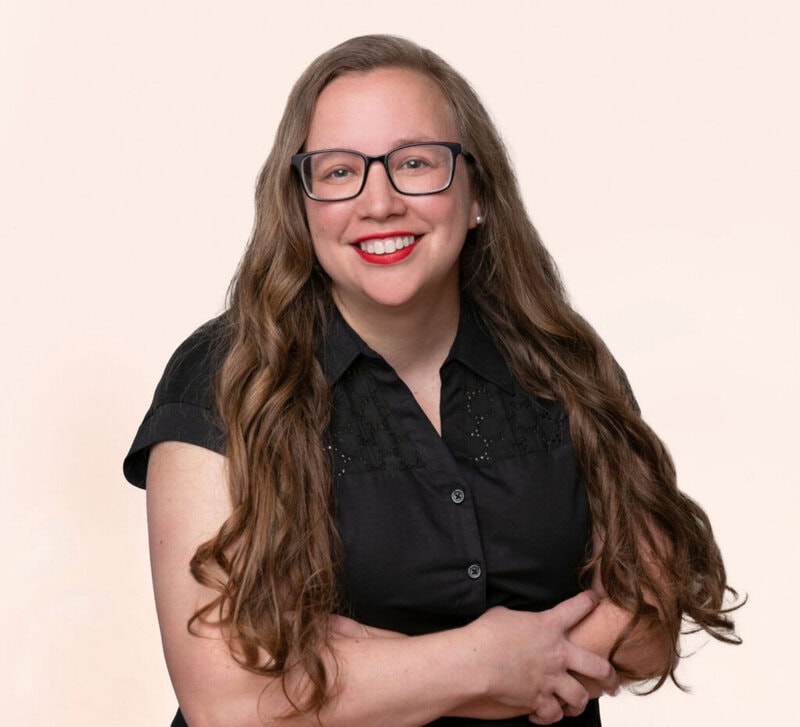 Woman with long brown hair and glasses, wearing a black short-sleeve shirt, smiling with arms crossed against a plain light background.