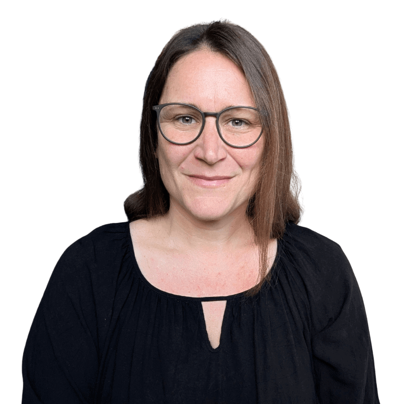 Goni Boller mit Brille und dunkler Bluse, lächelt vor hellgrauem Hintergrund in die Kamera