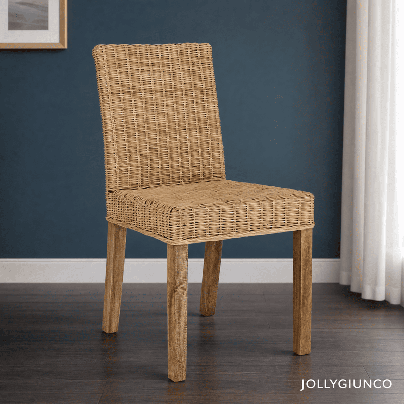 Wicker chair with wooden legs placed in a room with a dark blue wall; beige curtains to the right and a white baseboard visible; brand text in the bottom-right corner.