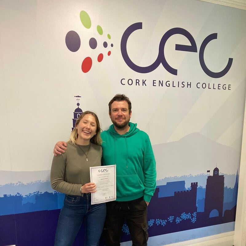 Glückliche Absolventin mit ihrem Zertifikat vor dem Logo des Cork English College.