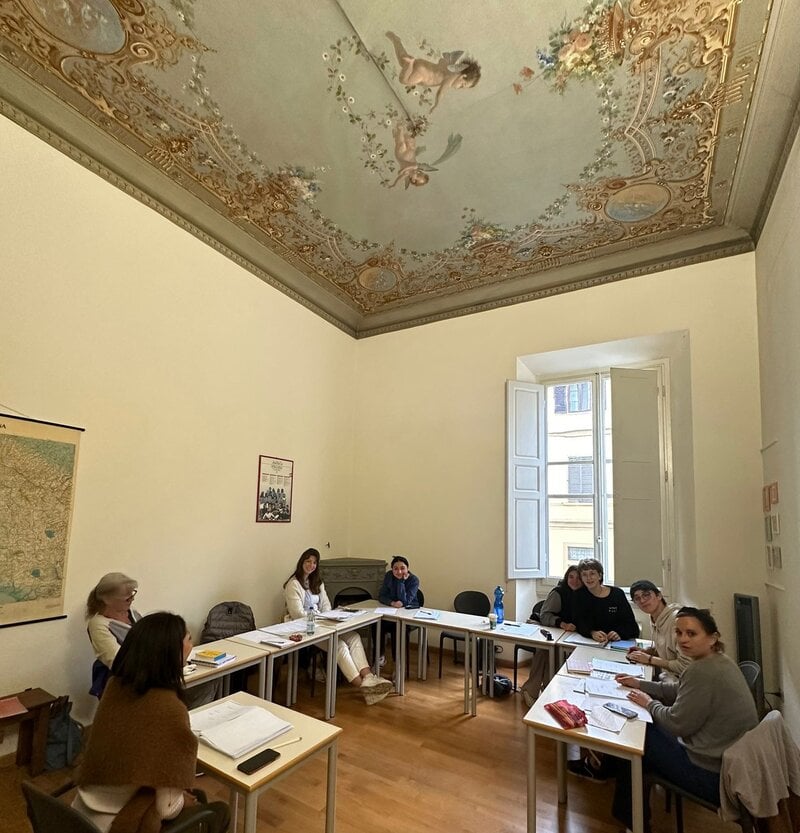 Ein historischer Klassenraum mit kunstvoll verziertem Deckenfresko, hohen Fenstern und einer Gruppe von Sprachschülern, die um einen Tisch sitzen.