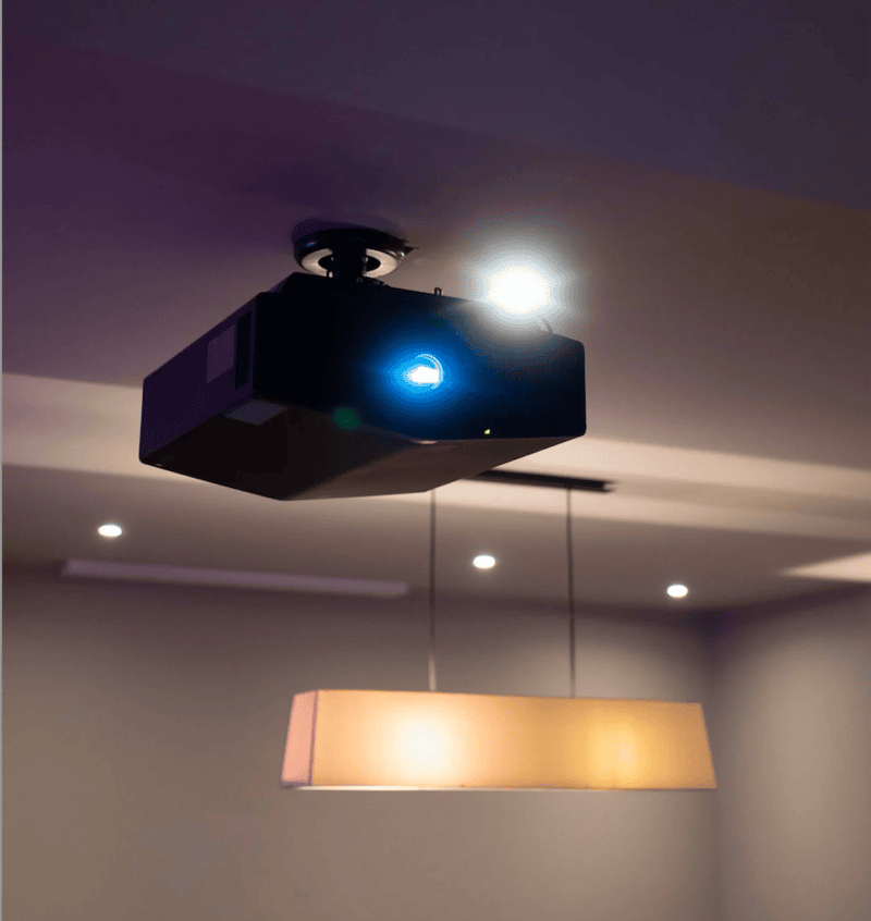 A ceiling-mounted projector is switched on, emitting a bright blue light in a dimly lit custom-built cinema room with pool table and bar in Fetcham, Surrey, featuring recessed ceiling lights and a rectangular pendant light hanging below.