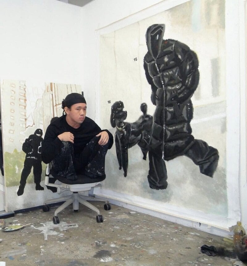 Yutaro Inagaki sitting in his studio beside a large painting of black puffer-clad humanoid figures.