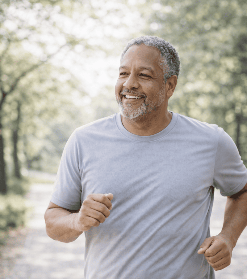 A middle-aged man in a gray T-shirt jogs outdoors on a tree-lined path, smiling, with sunlight filtering through the trees.
