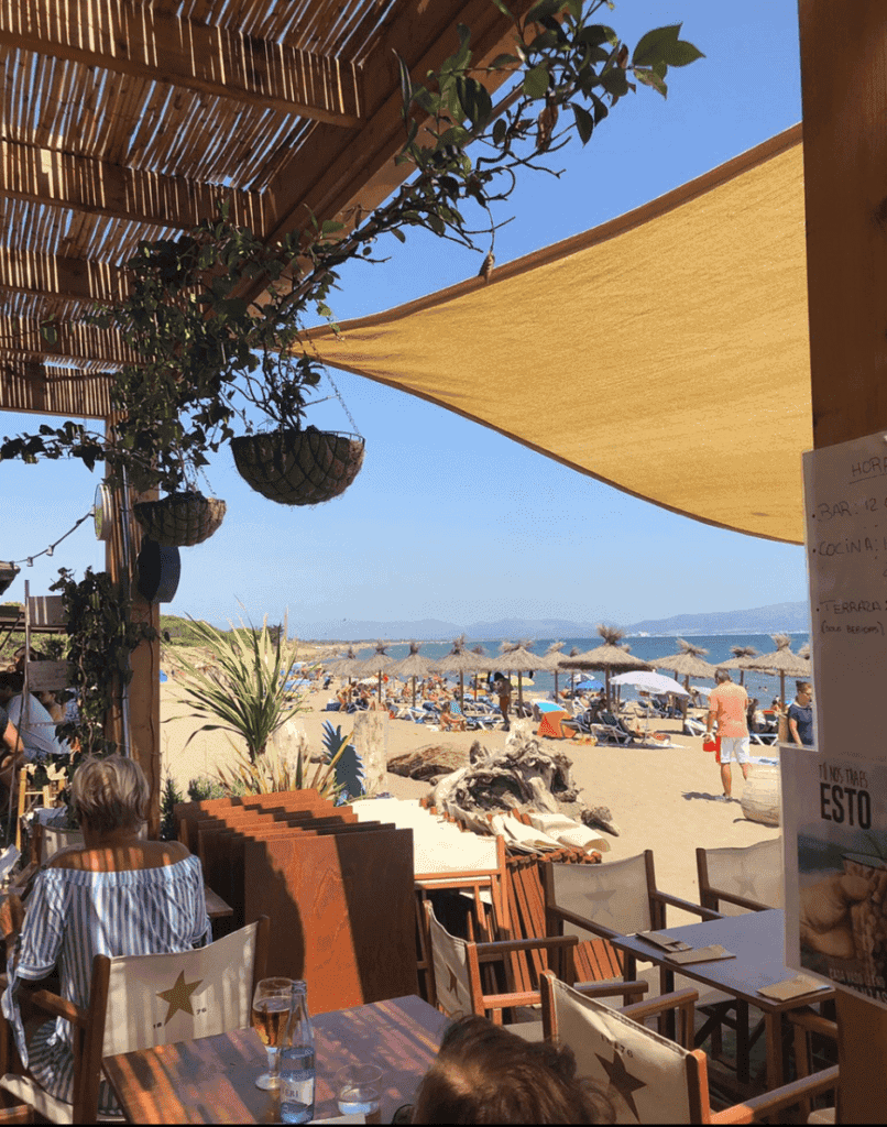 Beachside bar at can camades with outdoor seating, shaded umbrellas, and view of the sandy beach with sunbathers and thatched parasols, perfect for relaxing seaside dining and drinks in Mallorca, Spain.