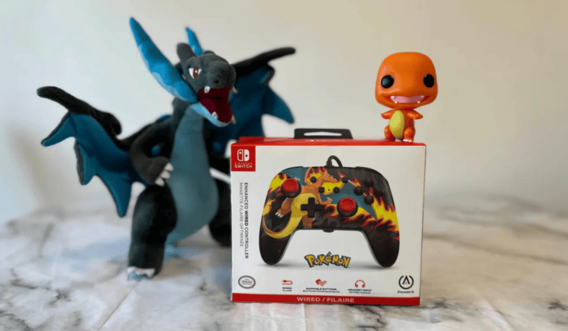 Photo of a boxed PowerA Enhanced Wired Controller Charizard Firestorm Edition. The box sits on a marble effect table. On top of the box is a little Charmander POP figure. By the side of it is a Mega Charizard X plushie toy.