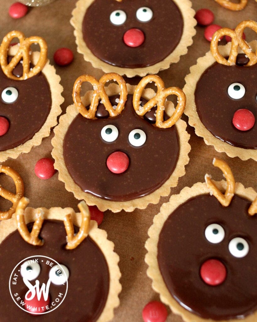 individual reindeer tarts on a greaseproof paper background. Chocolate tarts with pretzel antlers and red M&M noses and googly eyes