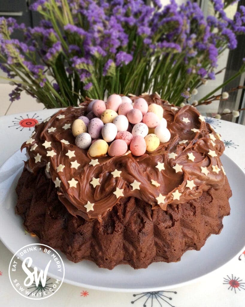 Chocolate Bundt Cake on a white table cloth with purple flowers behind.