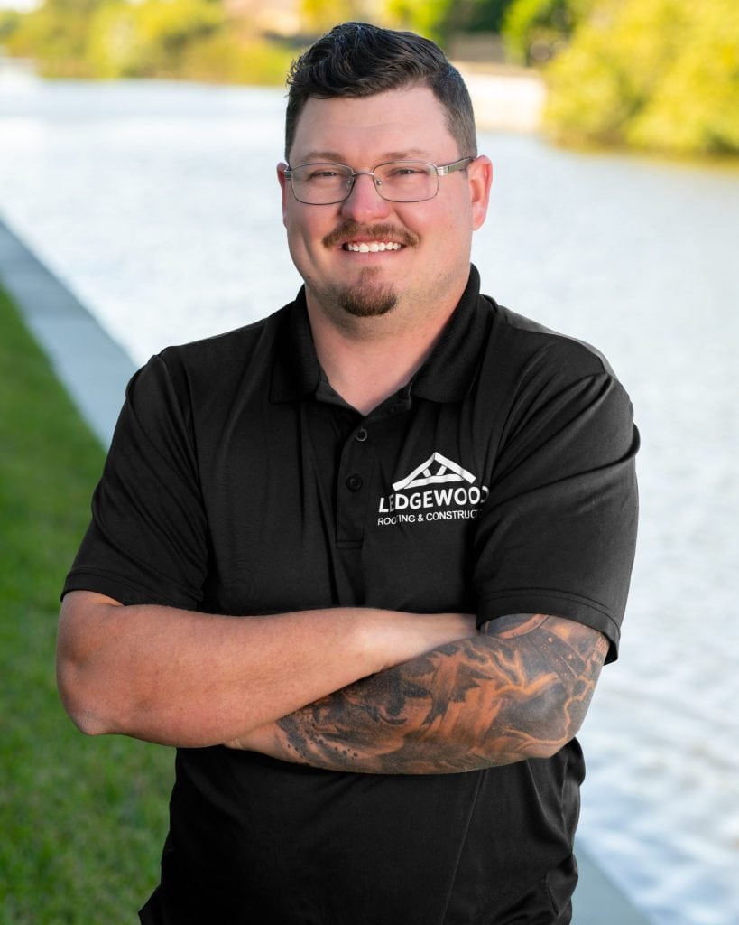 A man with glasses, short dark hair, and a mustache stands smiling with arms crossed near a canal. He wears a black Ledgewood Roofing polo and has a tattoo sleeve on his left arm. Trees and water complete the background.