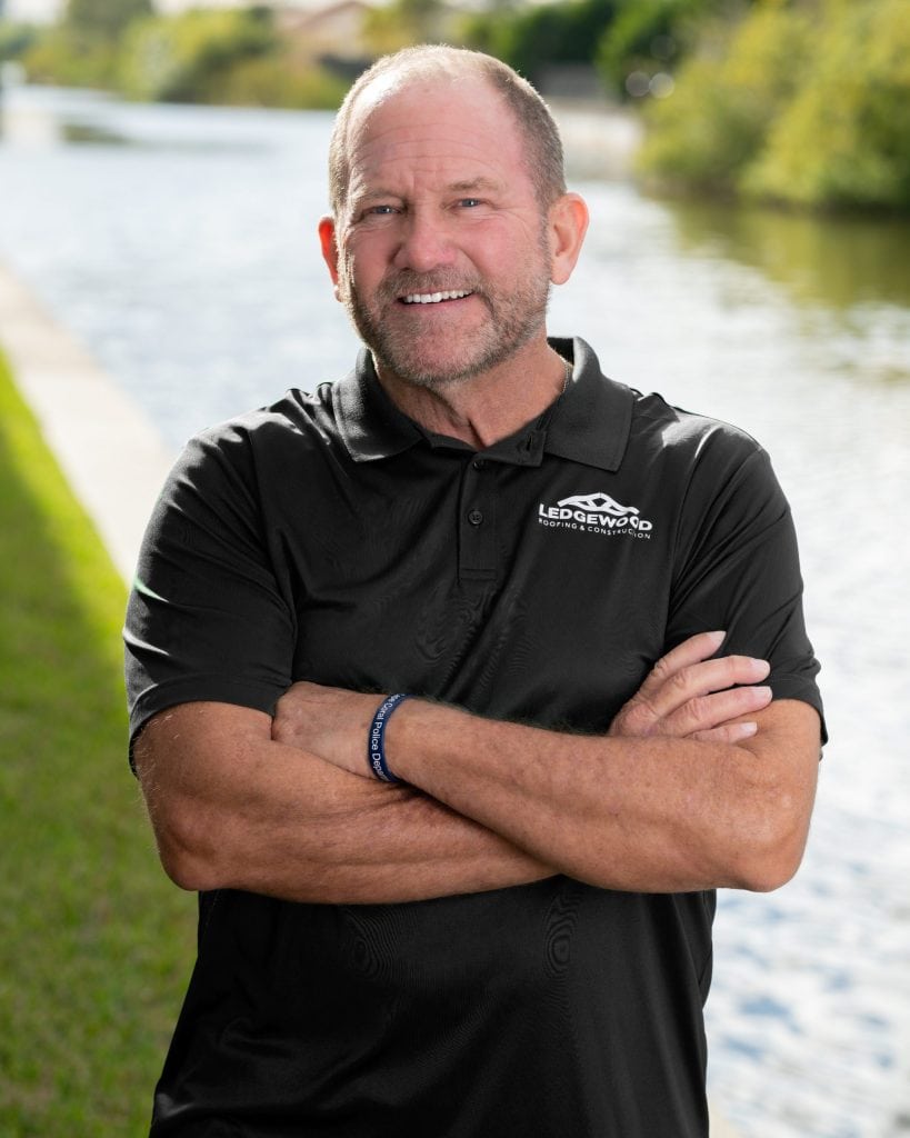 A man with short hair and a beard stands outdoors by a canal, smiling with arms crossed. He is wearing a black polo shirt with a Ledgewood Roofing logo and a blue wristband. Green grass and water are visible behind him.