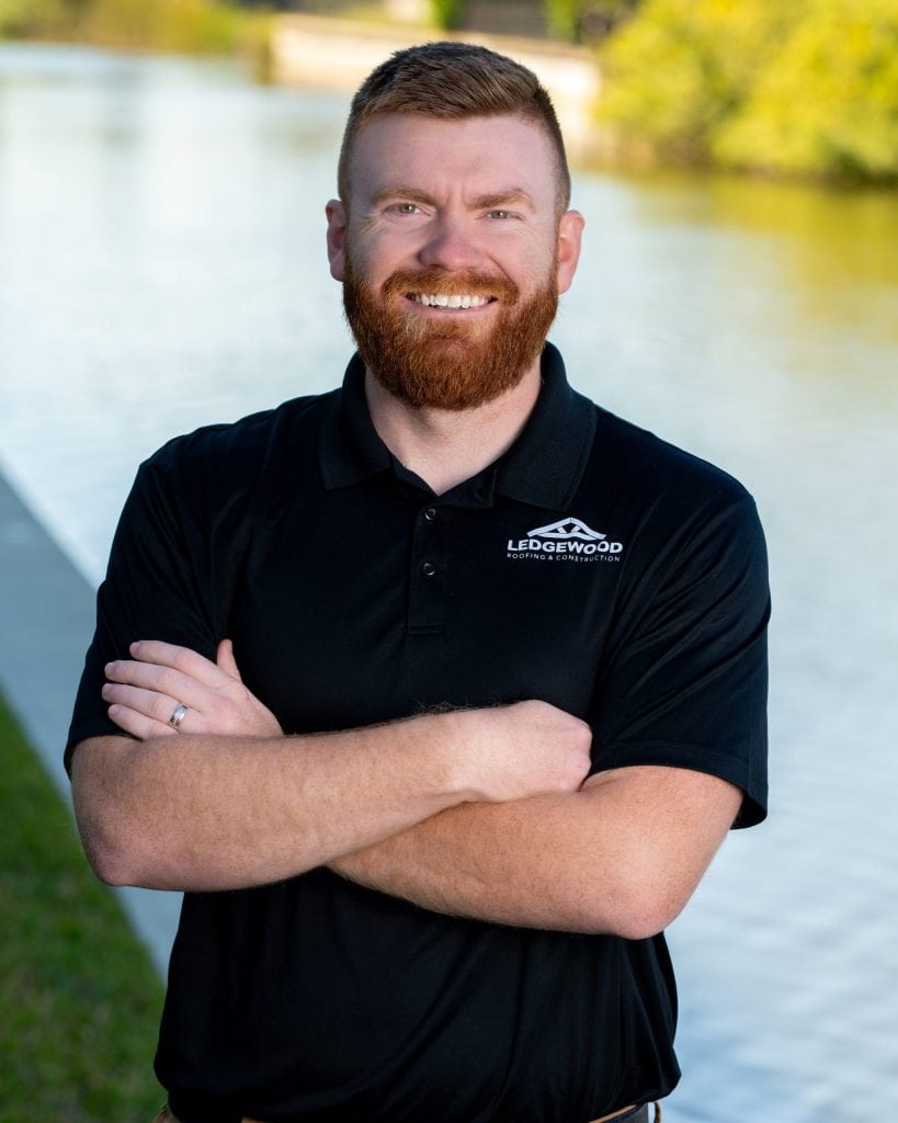 A smiling man with a red beard and short hair stands outdoors near water, wearing a black Ledgewood Roofing polo shirt and crossing his arms. Greenery and a concrete edge are visible in the background.