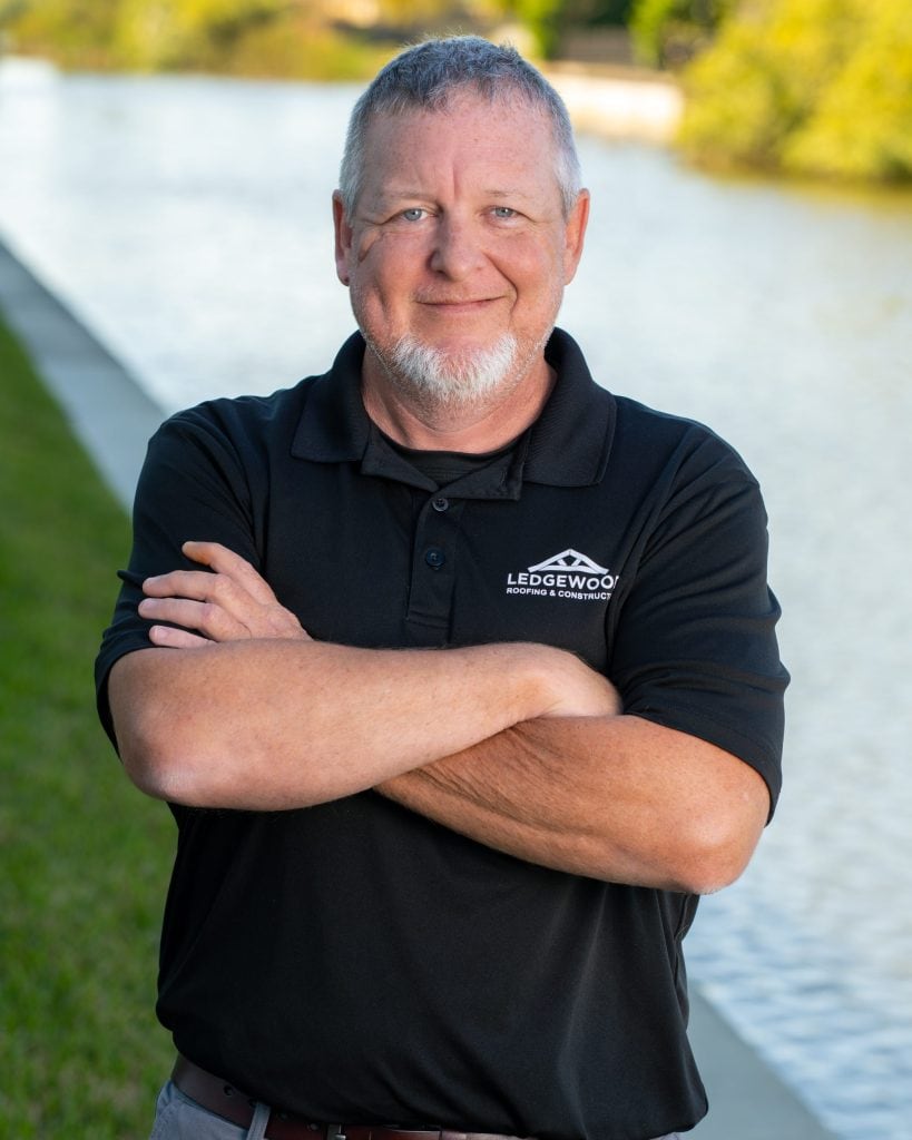 A man with short gray hair and a beard stands outdoors by a canal, smiling with arms crossed. He wears a black polo shirt featuring the Ledgewood Roofing logo. Green grass and water highlight the peaceful setting.