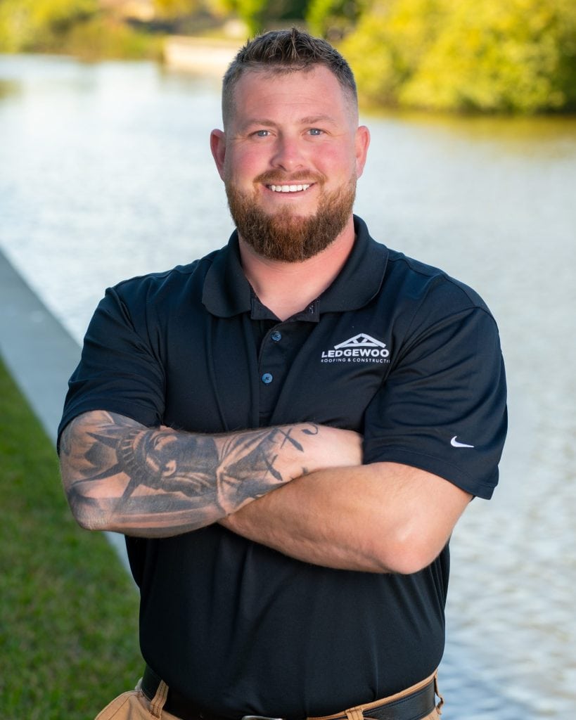A bearded man with tattooed arms stands smiling with his arms crossed, wearing a black Ledgewood Roofing and Construction polo shirt, outdoors by a waterway with greenery in the background.
