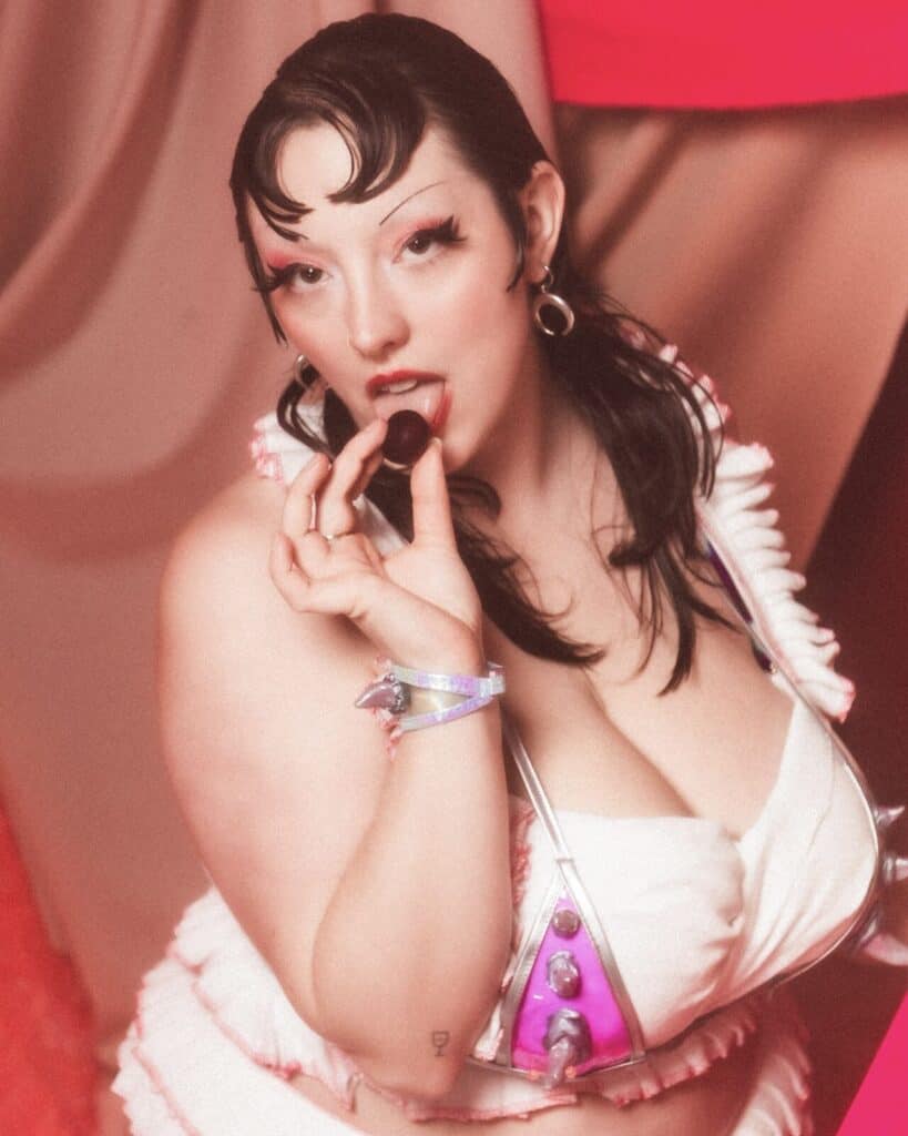A person with winged eyeliner and dark hair styled in waves wears a frilly white and pink outfit, holding a cherry to their lips, against a soft, warm-toned background.