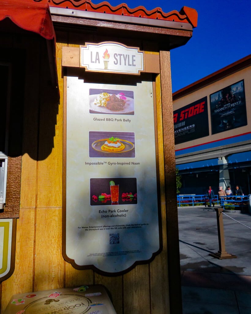 A 'LA Style' menu board at the Disney California Adventure Food and Wine Festival showcases delectable offerings under a sunny sky. In the background, the energetic ambiance of the park continues with guests and the 'Backlot Premiere Shop' sign visible.