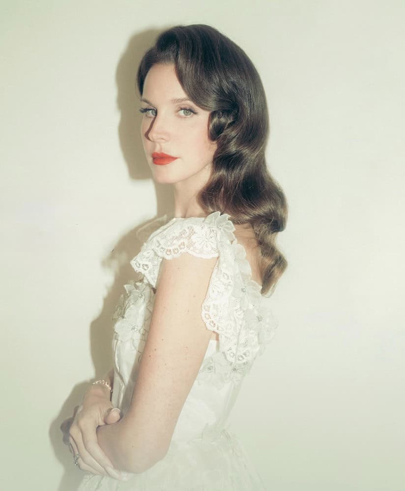 A woman with long wavy brown hair and red lipstick poses in a vintage-style white lace dress against a pale background, looking over her shoulder with a calm, elegant expression.