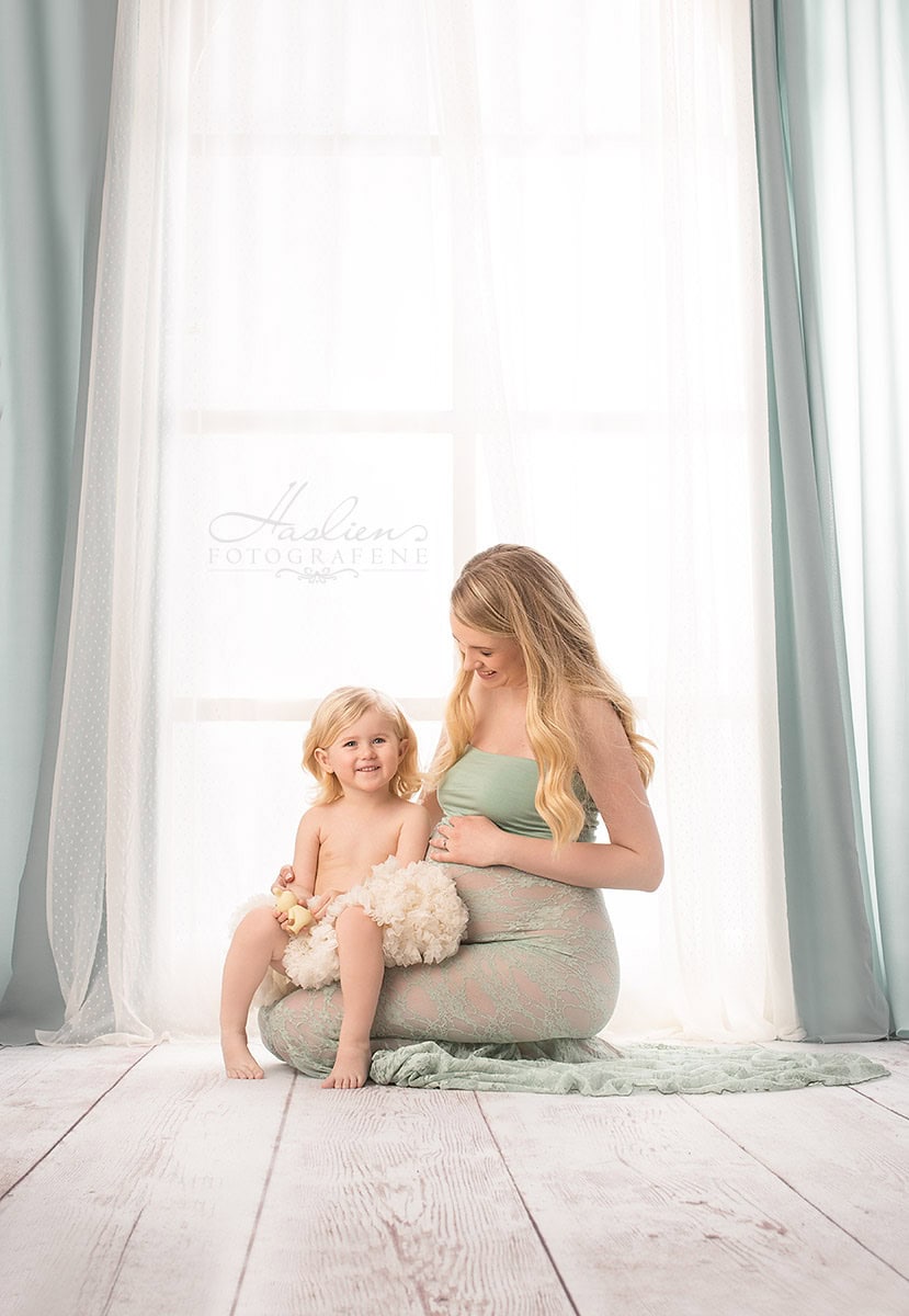 Livad gravidfoto med mor og liten jente foran stort vindu med lyse sommerfarger og myke gardiner. Nydelig gravidfotografering som fanger morens forventningsfulle øyeblikk og kjærligheten til det kommende barnet. En vakker gravidfotoopptak av mor og barn i intime omgivelser, der mor viser frem sin voksende mage. Perfekt for gravide fotosessioner, familiefotografering, og minnene om svangerskapet. relevans.