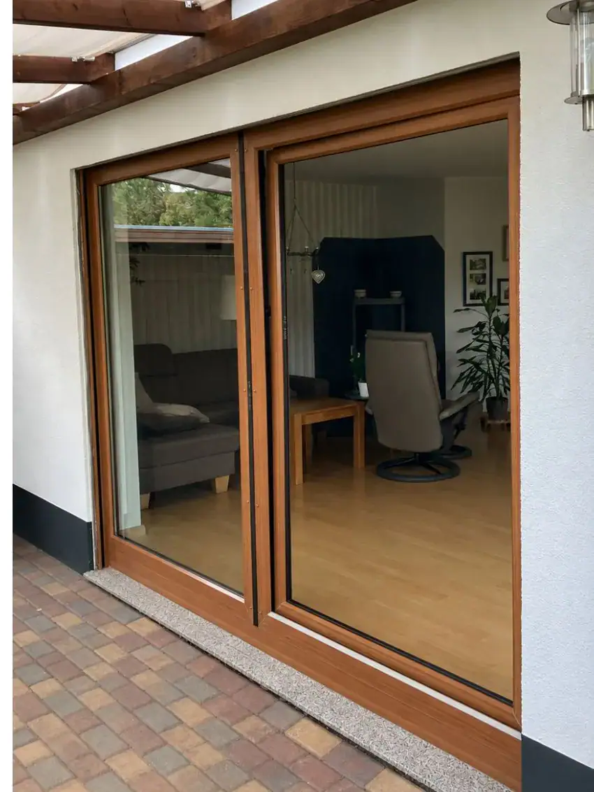 Braune Parallelschiebekipptür von Fütterer Fenster & Türen GmbH, die eine Verbindung zwischen einem gepflasterten Terrassenbereich und einem hellen Wohnraum mit Sofa und Holzboden bildet.