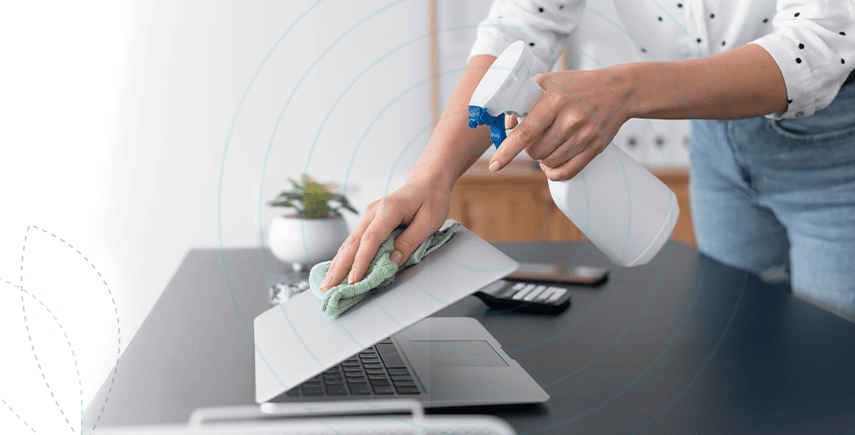 Person cleaning a laptop with a spray and cloth, using advanced hygiene solutions for a germ-free workspace.