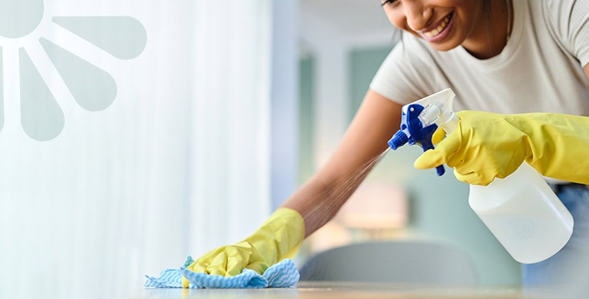 Smiling woman wearing yellow gloves cleaning a surface with a spray bottle and cloth in a bright room