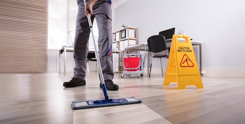 A janitor mopping the floor in a commercial office with a caution wet floor sign placed nearby.