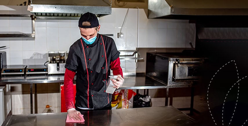 Chef in a commercial kitchen wearing a mask, cleaning a stainless steel surface with a cloth and sanitizer.