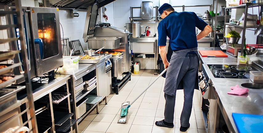 Chef mopping floor in a commercial kitchen with stainless steel equipment, ensuring hygiene and cleanliness.