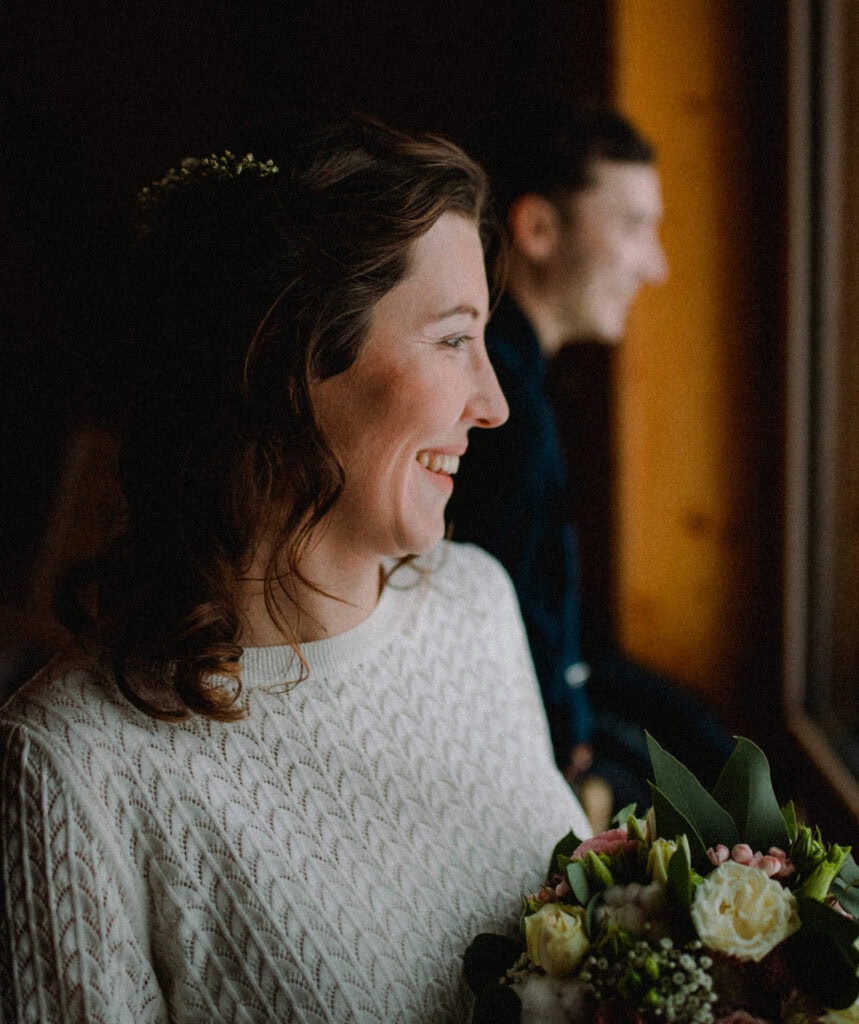 Eine Frau mit welligem Haar und cremefarbenem Pullover steht lächelnd neben einem Fenster und hält einen Blumenstrauß in der Hand. Die Szene wurde von einem Hochzeitsfotografen aus Regensburg aufgenommen und zeigt einen verschwommenen Mann im Hintergrund, der ebenfalls aus dem Fenster blickt.