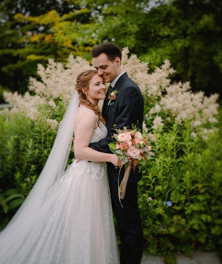 Braut und Bräutigam umarmen sich in einem üppigen Garten, aufgenommen von einem Hochzeitsfotografen aus Regensburg. Die Braut trägt ein fließendes weißes Kleid und hält einen Strauß rosa und pfirsichfarbener Blumen in der Hand, während der Bräutigam einen dunklen Anzug trägt. Umgeben von Grün und zarten, weiß blühenden Pflanzen fühlt sich ihr Moment zeitlos an.