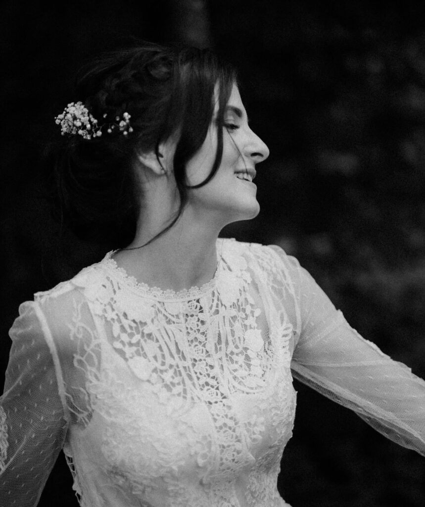 Schwarzweißfoto einer fröhlichen Frau in einem Spitzenbrautkleid, aufgenommen von einem Hochzeitsfotografen in Regensburg. Ihr Haar ist mit kleinen Blumen gestylt und sie lächelt, während sie vor einem dunklen, unscharfen Hintergrund zur Seite blickt.