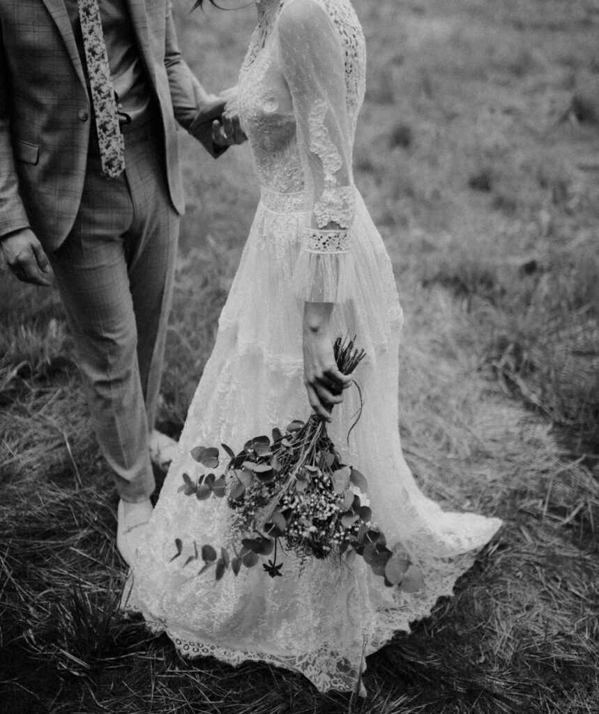 Das Schwarzweißfoto des Hochzeitsfotografen Regensburg zeigt eine Braut in einem Spitzenbrautkleid mit einem Blumenstrauß in der Hand, wie sie Hand in Hand mit dem Bräutigam durch eine Wiese geht.