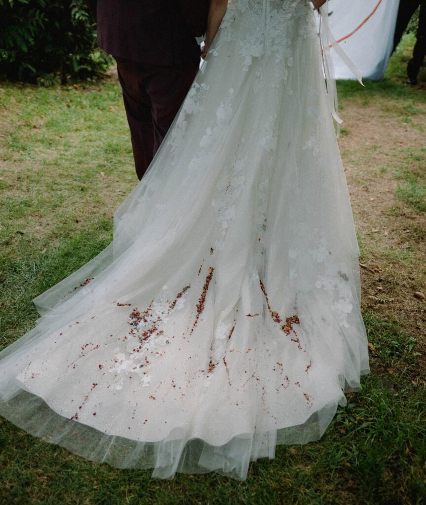 Das weiße Hochzeitskleid einer Braut, geschmückt mit roten Blütenblättern, liegt elegant auf dem grünen Rasen, während ein Hochzeitsfotograf in Regensburg den Moment festhält. Die Braut und eine weitere Person stehen zusammen, teilweise von hinten sichtbar, eingerahmt von majestätischen Bäumen und üppigem Laub.