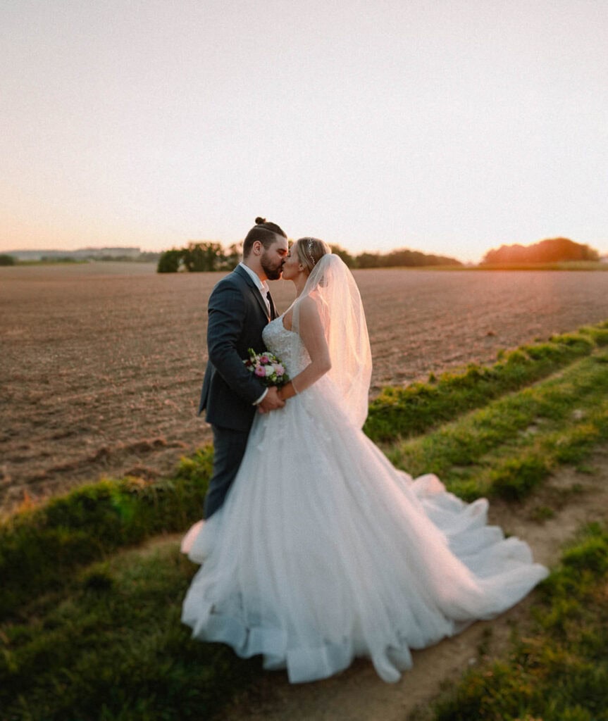 Braut und Bräutigam stehen bei Sonnenuntergang auf einem Feld, aufgenommen von einem Hochzeitsfotografen aus Regensburg. Die Braut trägt ein fließendes weißes Kleid und einen Schleier, während der Bräutigam einen dunklen Anzug trägt. Die Sonne wirft einen warmen Schein über die Landschaft und unterstreicht ihren intimen Moment.