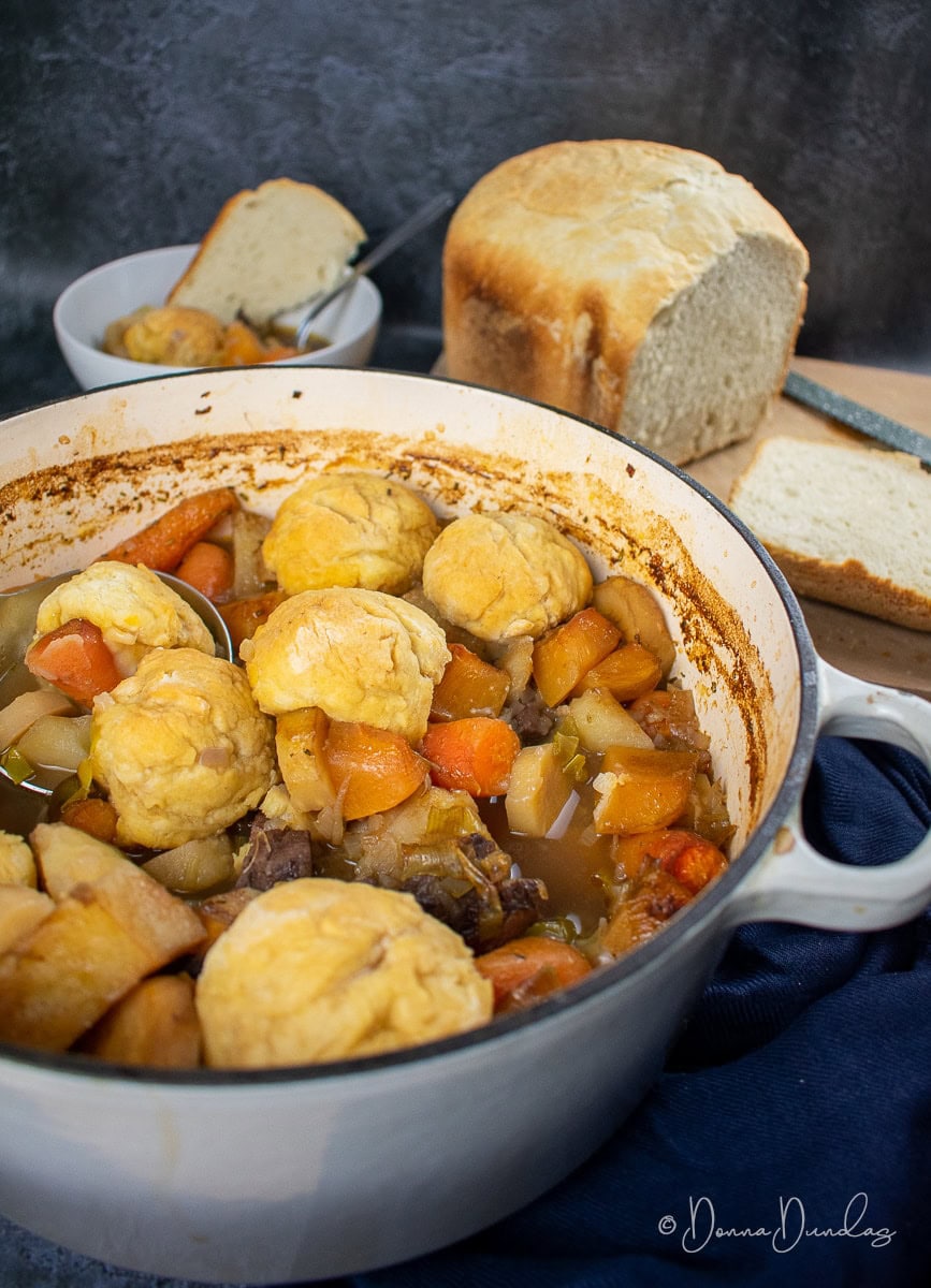 lamb stew with dumplings in cast iron pan, served portion in bowl and loaf of bread on chopping board in the background.