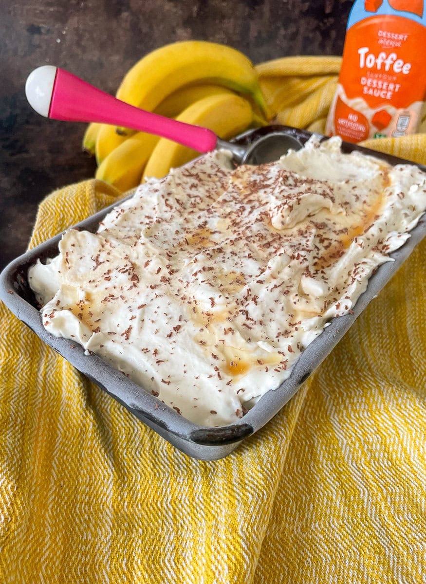 loaf tin of ice cream topped with toffee sauce and grated chocolate. Toffee sauce and bananas in the background.